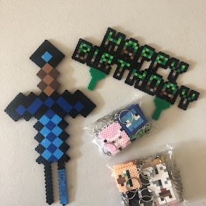 May include: A set of Minecraft-themed birthday party decorations, including a blue and brown pixelated sword, a green and brown pixelated banner that says "Happy Birthday", and a bag of pixelated keychains.