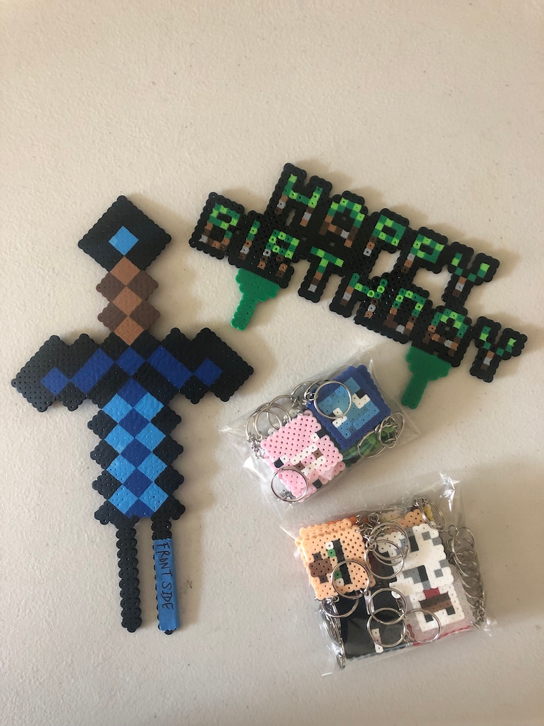 May include: A set of Minecraft-themed birthday party decorations, including a blue and brown pixelated sword, a green and brown pixelated banner that says "Happy Birthday", and a bag of pixelated keychains.