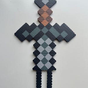 May include: A black, grey and brown pixelated sword made of beads. The sword has a diamond-shaped blade and a handle with a chequered pattern.
