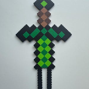 May include: A green and black pixelated sword made of plastic beads. The sword has a brown handle and a green blade.