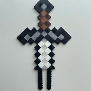 May include: A black and white pixel art sword made with perler beads. The sword has a brown handle and a grey blade.