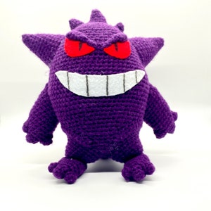 May include: Purple crocheted plush toy of a ghost Pokemon with red eyes and a white toothy grin.