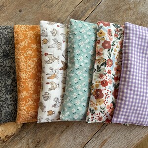 May include: Six rectangular fabric items with various patterns, including floral, chicken, and gingham designs. Colors include gray, orange, white, light blue, and purple. The items are arranged on a wooden surface.