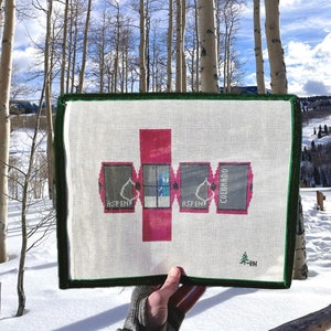 May include: Needlepoint canvas featuring a geometric design with the word "Aspen" and "Colorado". The canvas has a white background with a green border. The design is in shades of pink and gray. The canvas is held in front of a snowy landscape.