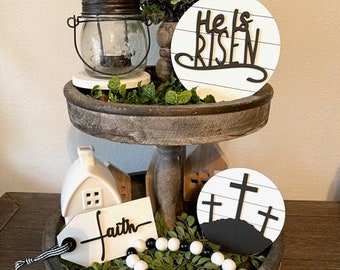 May include: A tiered tray decorated for Easter with a white sign that says "He is Risen", a white cross sign with the text "Blessed", a white sign with the text "Faith", a white sign with three crosses, and a beaded garland.