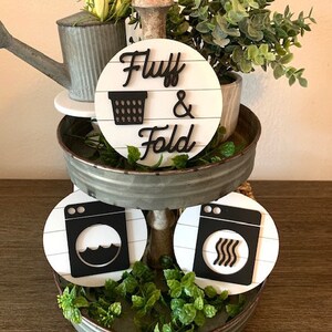 May include: A two-tiered tray with a white background and black text. The top tier has a sign that says "Fluff & Fold" with a black silhouette of a laundry basket. The bottom tier has two signs with black silhouettes of washing machines. The tray is decorated with greenery.