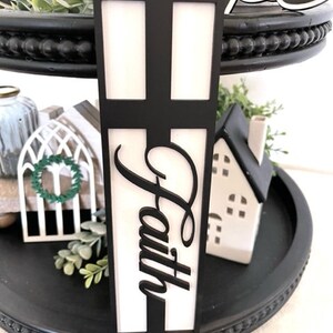Op de afbeelding: Een zwarte etag&egrave;re met twee niveaus met decoratieve items. Een zwart kruisvormig bord met het woord "Faith" in cursief is het middelpunt. Andere items zijn een klein wit huis, een decoratieve krans en groen.