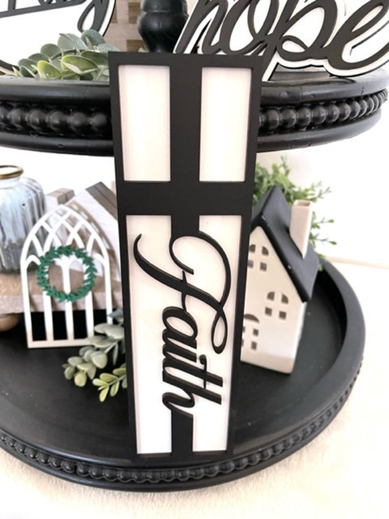 Op de afbeelding: Een zwarte etag&egrave;re met twee niveaus met decoratieve items. Een zwart kruisvormig bord met het woord "Faith" in cursief is het middelpunt. Andere items zijn een klein wit huis, een decoratieve krans en groen.