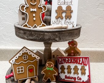 May include: A two-tiered wooden tiered tray decorated with gingerbread cookies, a gingerbread house, and signs that say "Baked with love" and "Gingerbread Baking Co."