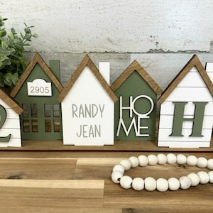May include: Wooden house-shaped decor with brown trim and white or green painted exteriors. Some houses display the number "2", the words "Randy Jean", "Home", and the letter "H". A decorative bead garland and greenery are also present.