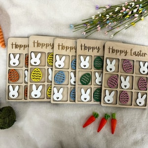 May include: Five wooden game boards with the text "Hoppy Easter" on one board and "Hoppy" on the other four boards. Each board has a grid of nine squares with bunny and egg cutouts.