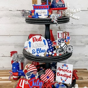 May include: A three-tiered tray decorated with red, white, and blue patriotic themed decorations for the 4th of July. The tray features a wooden sign that says "Uncle Sam's Fireworks", a red, white, and blue bicycle, a sign that says "Red White & Blue", and a sign that says "America Beautiful".