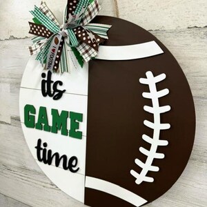 May include: A white and brown wooden door hanger shaped like a football. The football has white stitching and a white stripe across the middle. The left side of the door hanger has the words "it's game time" in green and black letters. A green, white, and brown bow is attached to the top of the door hanger.