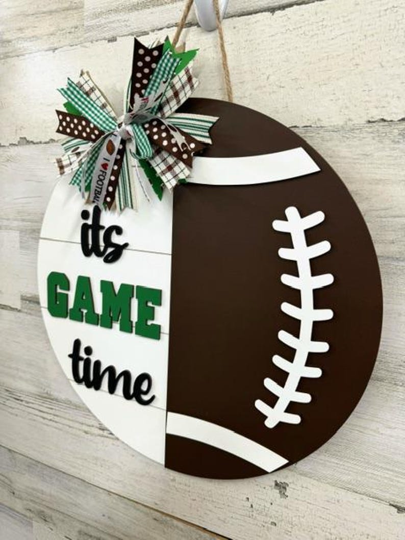 May include: A white and brown wooden door hanger shaped like a football. The football has white stitching and a white stripe across the middle. The left side of the door hanger has the words "it's game time" in green and black letters. A green, white, and brown bow is attached to the top of the door hanger.