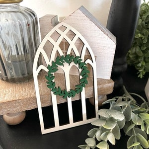 Op de afbeelding: Een decoratief houten boogvenster met een groene krans in het midden. Het raam is wit met een traliewerkontwerp. Een heldere glazen pot met een gouden deksel staat links van het raam. Het raam staat op een houten standaard.
