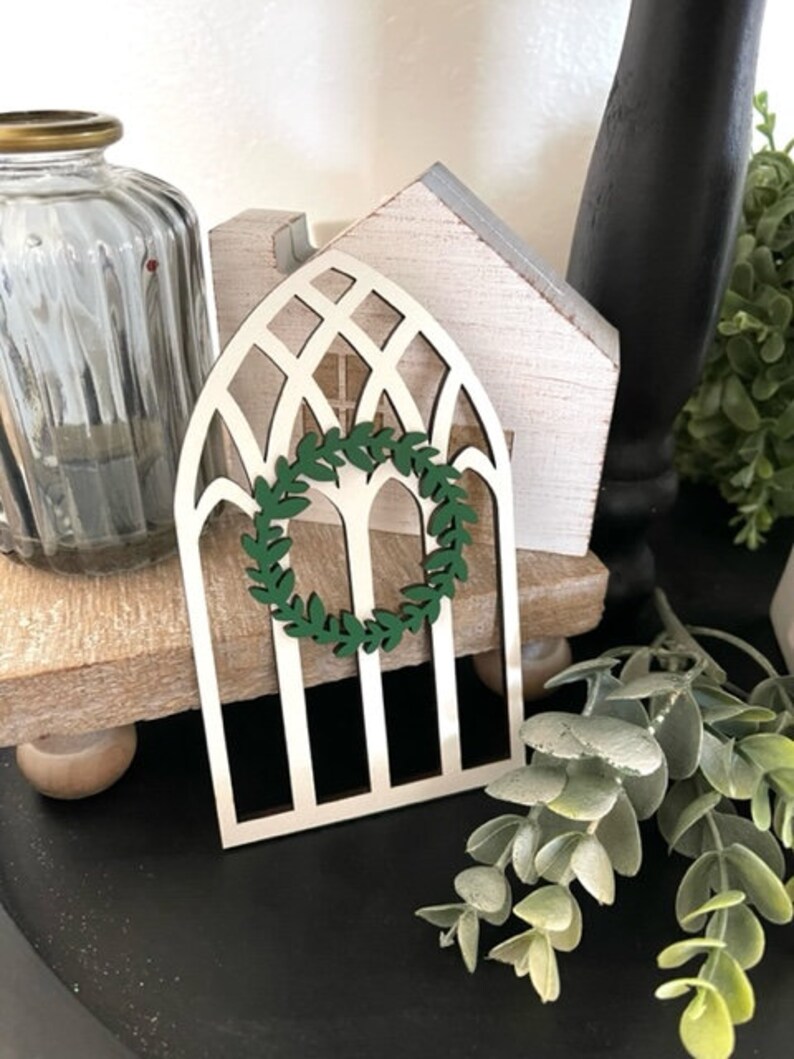 Op de afbeelding: Een decoratief houten boogvenster met een groene krans in het midden. Het raam is wit met een traliewerkontwerp. Een heldere glazen pot met een gouden deksel staat links van het raam. Het raam staat op een houten standaard.