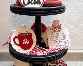 May include: A three-tiered black tiered tray decorated with a snowman, a small Christmas tree, and various red and white Christmas-themed decorations. The tray features the words "Hot Cocoa", "Warm & Cozy", "Have a Cup of Cheer", and a menu listing "Hot Cocoa, Marshmallows, Whipped Cream, Candy Canes, Sprinkles & Cheer".