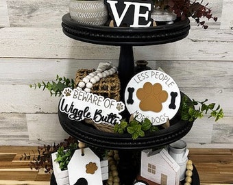 May include: A three-tiered black tiered tray decorated with dog-themed wooden signs, a white ceramic house, a white ceramic dog tag, a brown wicker basket, and a variety of greenery. The signs read "Love", "Beware of Wiggle Butts", "Less People More Dogs", and "The Sassy Designs".