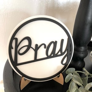 Op de afbeelding: Een ronde, witte plaat met het woord "Pray" in zwart geschreven. De plaat heeft een zwarte rand en staat op een kleine houten standaard. De achtergrond is een wazige afbeelding van een zwart oppervlak en groen.