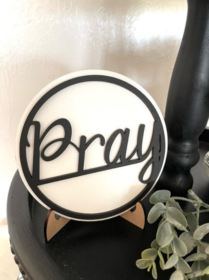 Op de afbeelding: Een ronde, witte plaat met het woord "Pray" in zwart geschreven. De plaat heeft een zwarte rand en staat op een kleine houten standaard. De achtergrond is een wazige afbeelding van een zwart oppervlak en groen.