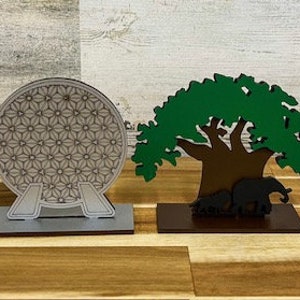 May include: Four wooden Disney park icon ornaments. The icons are Mickey Mouse's head, the Spaceship Earth sphere, the Tree of Life, and Cinderella's Castle. The ornaments are on a wooden surface.