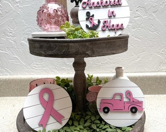 May include: Two-tiered wooden tray with pink and white decorations. The top tier has a pink and white sign that says "Anchored in Hope" with a pink ribbon anchor. The bottom tier has two pink and white signs, one with a pink ribbon and the other with a pink truck.