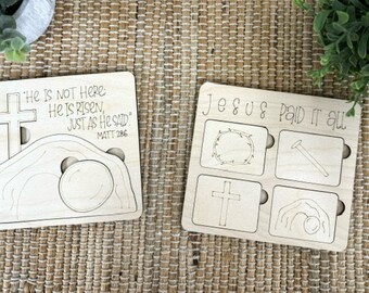 May include: Two wooden Easter puzzle pieces. The first piece has a picture of a tomb with a cross and the text "He is not here: He is risen, just as He said. Matt 286". The second piece has a picture of a crown of thorns, a nail, a cross, and a tomb with a rolling stone. The text "Jesus Paid It All" is at the top of the second piece.