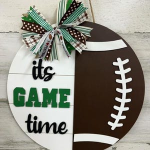 May include: A white and brown football-shaped door hanger with the text "it's game time" in black and green letters. A green, white, and brown bow is attached to the top of the hanger.
