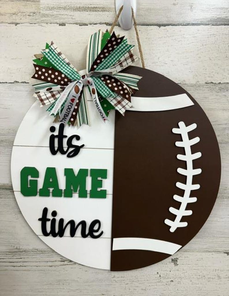 May include: A white and brown football-shaped door hanger with the text "it's game time" in black and green letters. A green, white, and brown bow is attached to the top of the hanger.