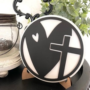 Op de afbeelding: Een zwart-wit decoratief rond bord met een hart- en kruisontwerp. Het bord wordt weergegeven op een kleine houten standaard, met een decoratieve handgreep en groen in de achtergrond. Het is een religieus thema.