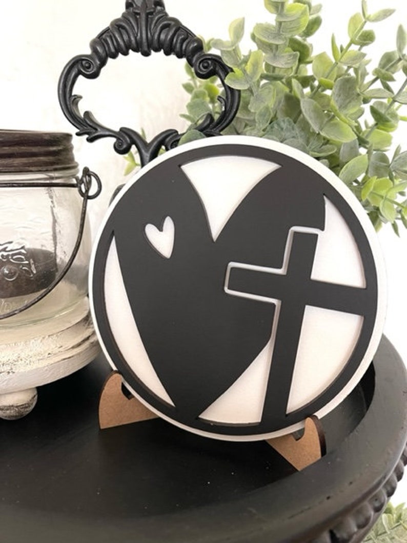 Op de afbeelding: Een zwart-wit decoratief rond bord met een hart- en kruisontwerp. Het bord wordt weergegeven op een kleine houten standaard, met een decoratieve handgreep en groen in de achtergrond. Het is een religieus thema.
