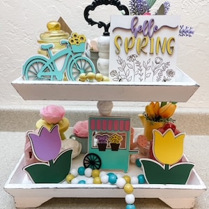 May include: A white tiered tray decorated with spring-themed wooden cutouts. The top tier features a turquoise bicycle with yellow flowers, a glass jar, and a sign that reads "hello spring". The bottom tier has a turquoise flower stand with pink and yellow flowers, a purple tulip, a yellow tulip, and a white and blue beaded garland.