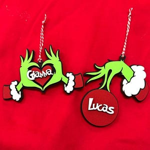 May include: Two hanging ornaments featuring green cartoon hands with white fur cuffs. One ornament shows hands forming a heart shape around the name Gianna in white text. The other shows a hand holding a red circular disc with the name Lucas in white text.