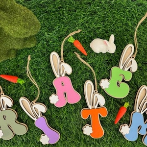 May include: Eight wooden Easter bunny ornaments, each shaped like a letter of the alphabet, painted in different colors. The letters are A, E, R, T, M, and H. The ornaments are hanging from twine and have a white bunny tail.