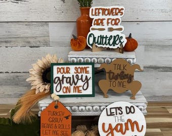 Könnte beinhalten: Dekorative Holzschilder und Akzente zum Thema Thanksgiving. Enthält eine Vase mit Grün, Kürbissen, einer Sonnenblume und Schilder mit Sätzen wie "Pour some gravy on me" und "Let's do the yam thing." Die Farbpalette umfasst Orange, Braun, Grün und Weiß.