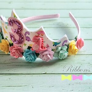 May include: A white headband with a pink, white, and green floral design. The headband has a pink number three with glitter and a pink dinosaur on it. The headband says "Ribbons Room" on the bottom.