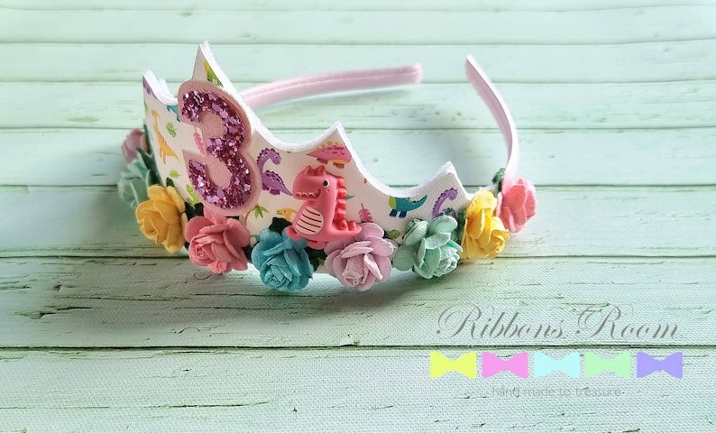 May include: A white headband with a pink, white, and green floral design. The headband has a pink number three with glitter and a pink dinosaur on it. The headband says "Ribbons Room" on the bottom.