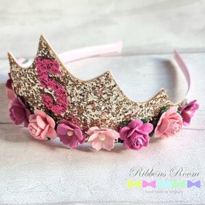 May include: Gold glitter crown headband with pink felt flowers and a pink felt number 5. The headband is decorated with gold glitter and pink felt flowers. The number 5 is made of pink felt and is attached to the crown.