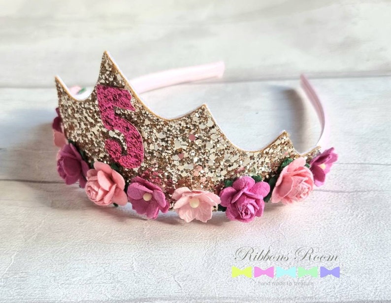 May include: Gold glitter crown headband with pink felt flowers and a pink felt number 5. The headband is decorated with gold glitter and pink felt flowers. The number 5 is made of pink felt and is attached to the crown.