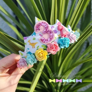 May include: A white headband with a pink glitter number 3, dinosaur print, and colorful fabric flowers. The headband is decorated with pink, blue, yellow, and green flowers.