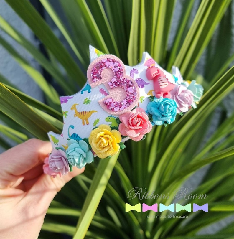 May include: A white headband with a pink glitter number 3, dinosaur print, and colorful fabric flowers. The headband is decorated with pink, blue, yellow, and green flowers.