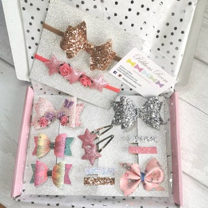 May include: A collection of glittery hair accessories, including bows, star-shaped clips, and headbands, presented in a pink box with a silver glitter base. The accessories feature various colors and textures, with a focus on pink, gold, and silver.