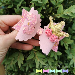 K&ouml;nnte beinhalten: Zwei rosa Haarschleifen mit Glitzerakzenten und einem gewellten Rand. Eine hat eine glitzernde "3", die andere eine goldene Krone und rosa Rosen. Die Schleifen sind vor gr&uuml;nem Laub platziert. Der Text "Ribbons Room" ist sichtbar.
