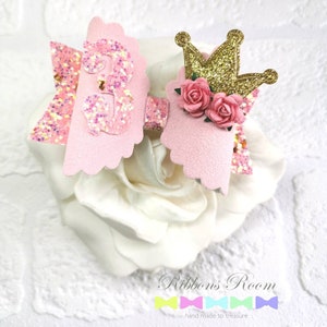 K&ouml;nnte beinhalten: Eine rosa-goldene Haarschleife mit einer glitzernden Zahl 3, einer goldenen Krone und rosa Rosen. Die Schleife ist auf einer wei&szlig;en Blume platziert. Der Text "Ribbons' Room" und "hand made to treasure" befinden sich unten.