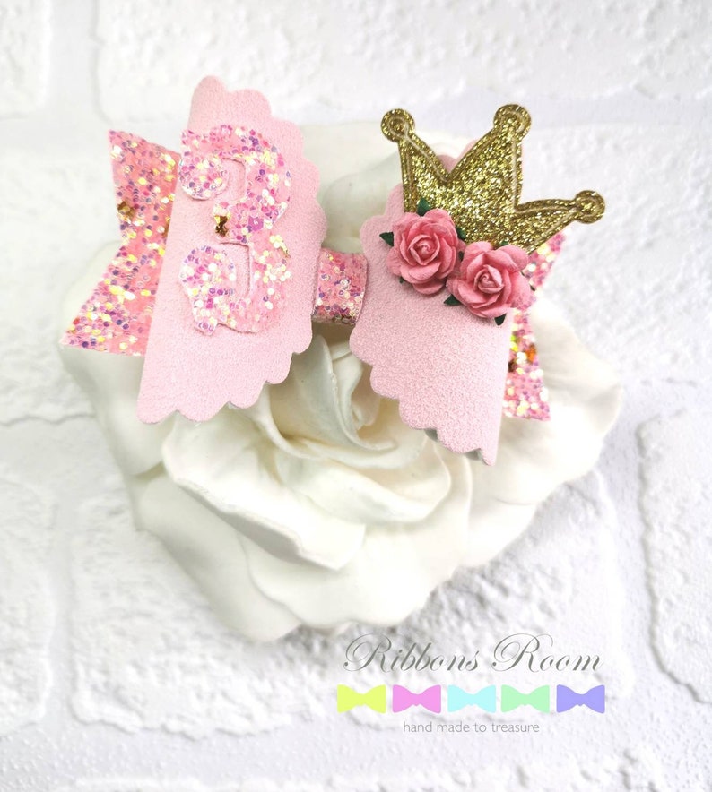 K&ouml;nnte beinhalten: Eine rosa-goldene Haarschleife mit einer glitzernden Zahl 3, einer goldenen Krone und rosa Rosen. Die Schleife ist auf einer wei&szlig;en Blume platziert. Der Text "Ribbons' Room" und "hand made to treasure" befinden sich unten.