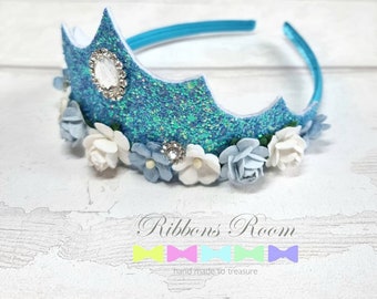 May include: A glittery blue crown with a turquoise headband, adorned with white and blue fabric flowers and sparkling embellishments. The crown features a decorative design and the text "Ribbons Room".
