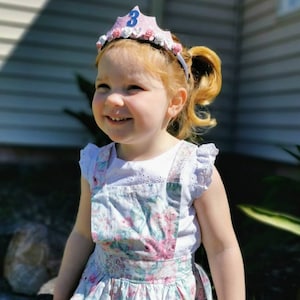May include: A young girl wearing a pink and white crown with a blue number 3 on top. She is wearing a white shirt with a floral print overall dress.