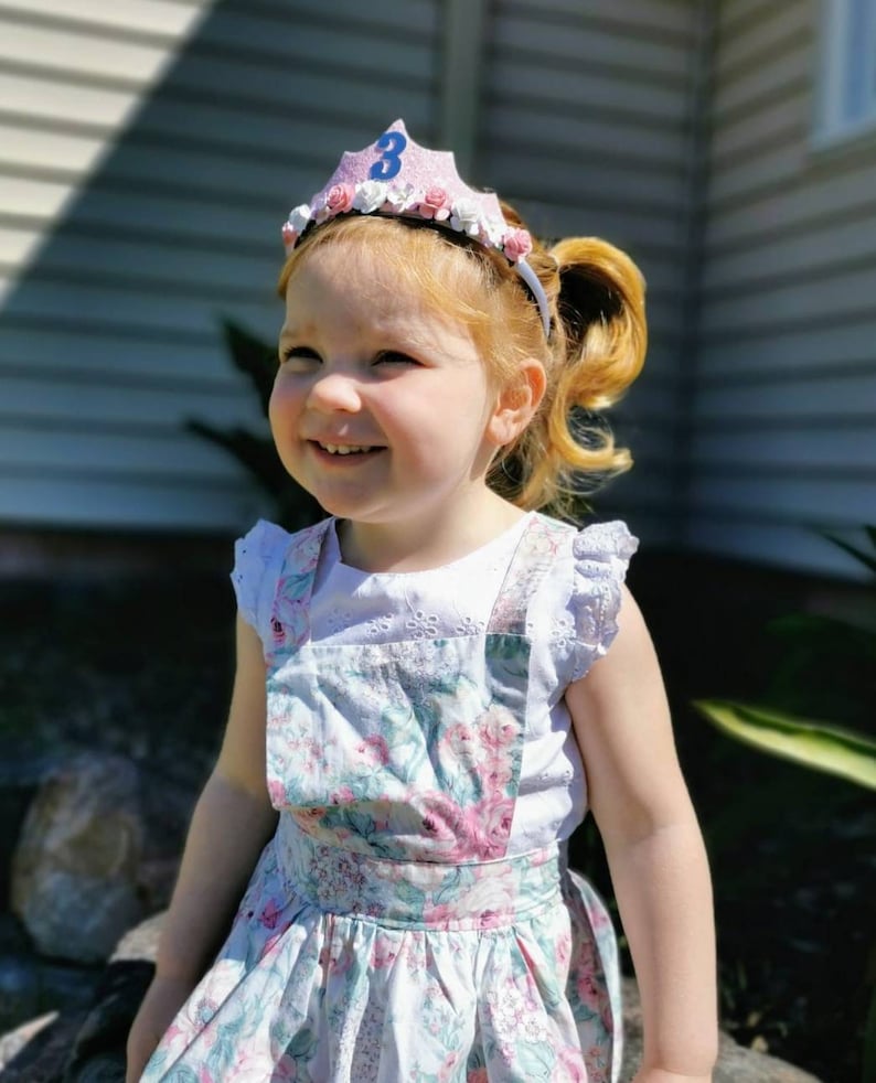 May include: A young girl wearing a pink and white crown with a blue number 3 on top. She is wearing a white shirt with a floral print overall dress.