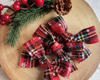 May include: Three red plaid bows with white snowflake accents are arranged on a wooden surface, alongside a sprig of evergreen, red berries, and a pinecone. The bows have a festive, holiday theme.