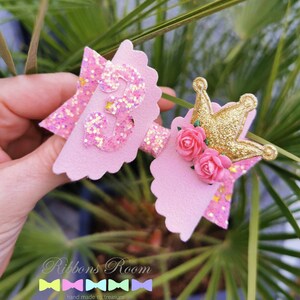 K&ouml;nnte beinhalten: Zwei rosa glitzernde Haarschleifen mit einem gewellten Rand. Eine Schleife hat die Zahl "3" in Glitzer, die andere eine goldene Glitzerkrone und rosa Rosen. Die Schleifen sind vor gr&uuml;nem Laub angeordnet.